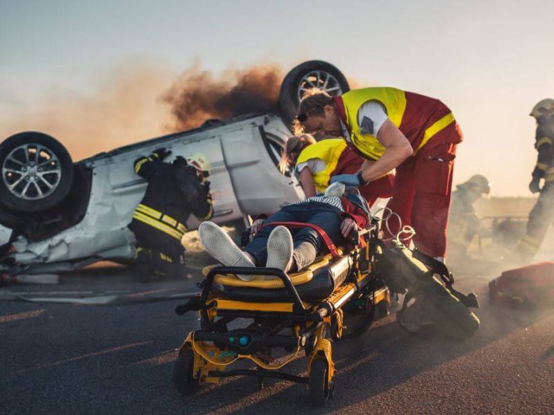A paramedic attends to an injured person on a stretcher against the backdrop of an overturned, smoking vehicle with responders nearby.