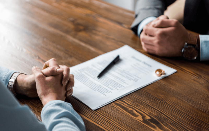 A document with text lies on a wooden table, while two hands clasped in contemplation face each other, suggesting a serious discussion or negotiation in a professional setting. A pen rests nearby.