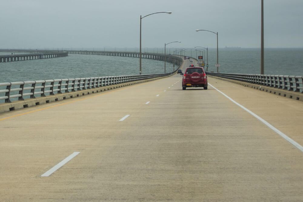 A view of the Hampton Roads Bridge-Tunnel with a red car driving along the bridge section on an overcast day. The bridge spans across a wide body of water, featuring safety barriers on either side and multiple lanes marked by clear white lines. In the distance, a parallel bridge section can be seen running alongside.