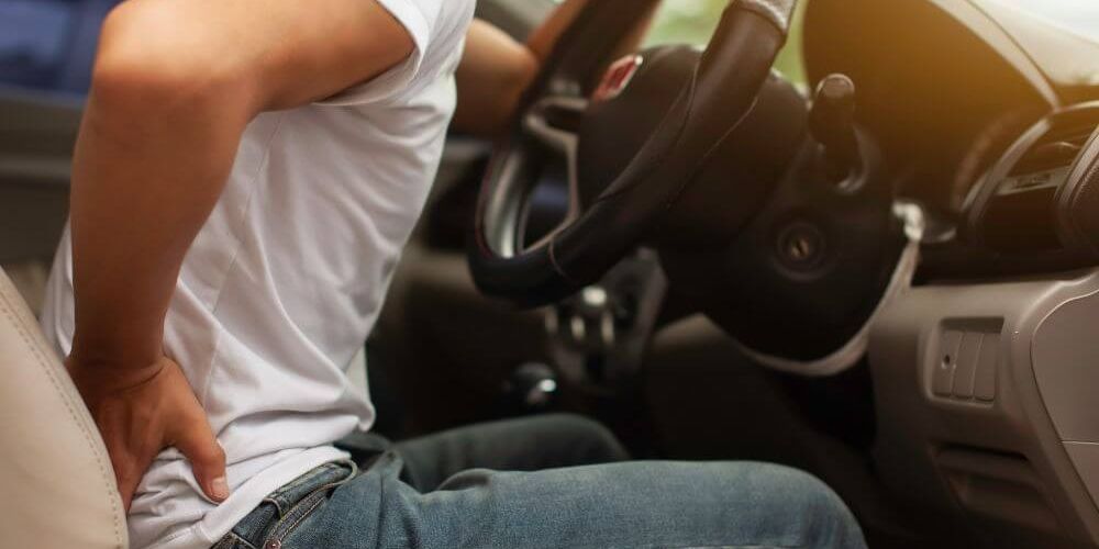 A person in a white shirt sits in a car, gripping the steering wheel while wincing and holding their lower back, suggesting discomfort in the vehicle's interior.