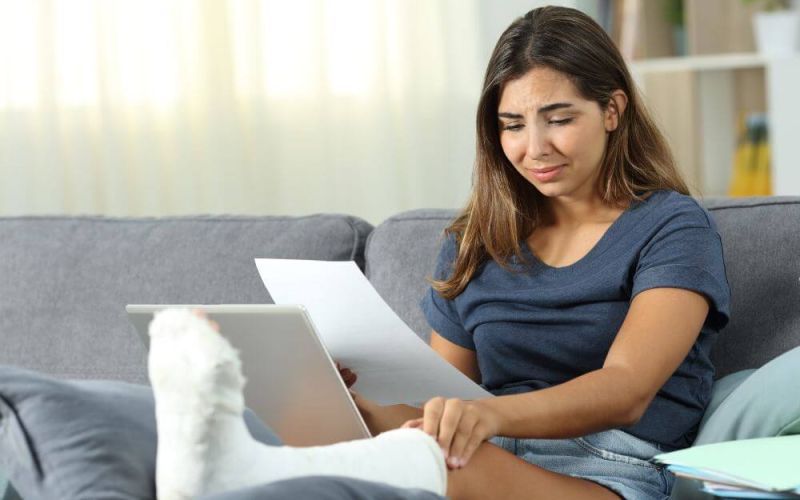 Woman with a leg cast sitting on a sofa, reading a document, looking concerned.