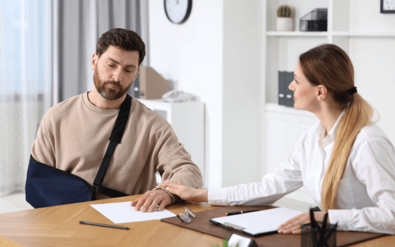 A man with an arm sling sits at a table with a woman in a white coat, who is gently holding his hand. They are in an office with shelves and a clock.