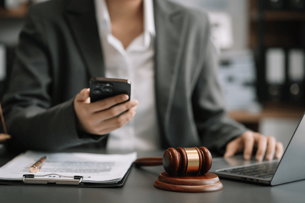 A person in a suit multitasks with a smartphone and laptop, near a gavel on a desk, possibly suggesting a legal setting.