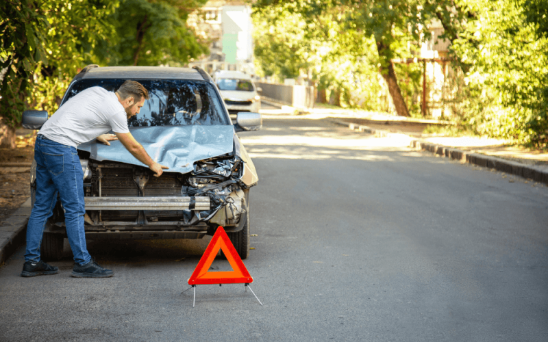 A man inspects a damaged car hood on a quiet, tree-lined street. A red warning triangle is placed on the road in front of the vehicle.