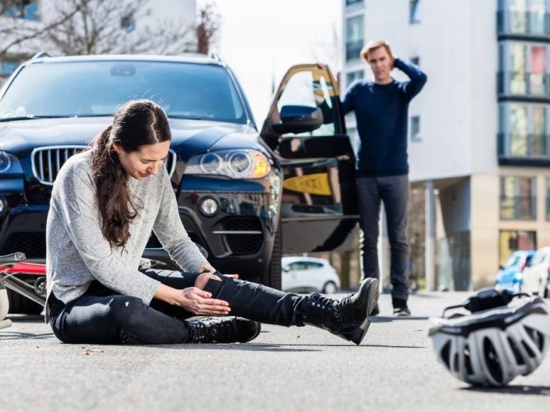 Pedestrian sitting injured on road after car accident, driver concerned, bike helmet nearby.