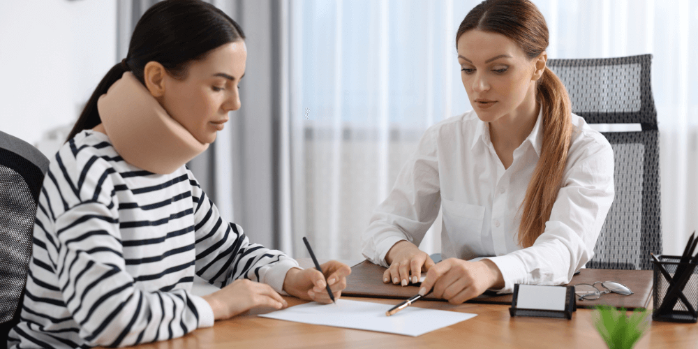 A woman with a neck brace sits at a table, writing while another woman in a white shirt points to paperwork, both engaged in discussion in a bright, modern office setting.