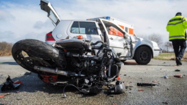 A damaged motorcycle lies on the road in front of a white car with severe side damage. The scene appears to be an accident, with a person in a yellow reflective jacket standing nearby. The background shows a cloudy sky and an open area, suggesting a roadside location in Virginia.