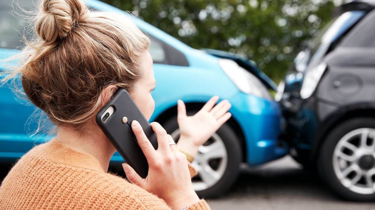 A woman on a phone gestures in frustration near two cars that have collided, emphasizing a car accident scenario.