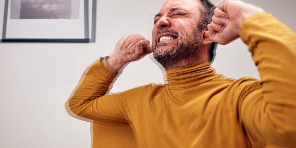 A man in a yellow turtleneck sits on a couch, grimacing and covering his ears with his hands, clearly distressed. A black-and-white photo hangs on the wall behind him.