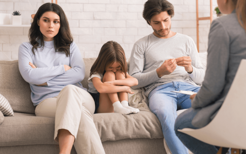 A family sits on a couch during a counseling session; a woman looks upset, a man appears contemplative, and a child rests her head on her knees; a therapist takes notes nearby.