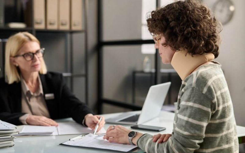 Two people sit at a desk, one signing a document; in an office with shelves and a laptop, suggesting a professional setting.