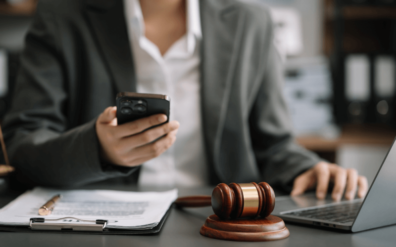 A person in a suit multitasks with a smartphone and laptop, near a gavel on a desk, possibly suggesting a legal setting.