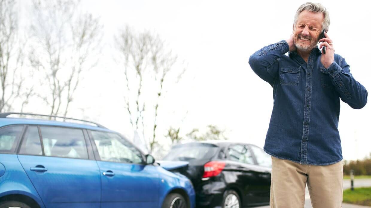 A distressed senior man standing next to his blue car after a minor traffic collision, talking on his mobile phone. He appears frustrated and is holding his head with one hand while speaking. The setting is a roadside with leafless trees in the background, suggesting a cold or transitional season. This image could represent a scenario where someone might need the services of a personal injury lawyer in Virginia Beach.