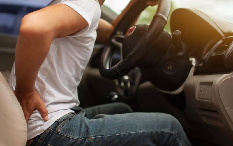 A person in a white shirt sits in a car, gripping the steering wheel while wincing and holding their lower back, suggesting discomfort in the vehicle's interior.