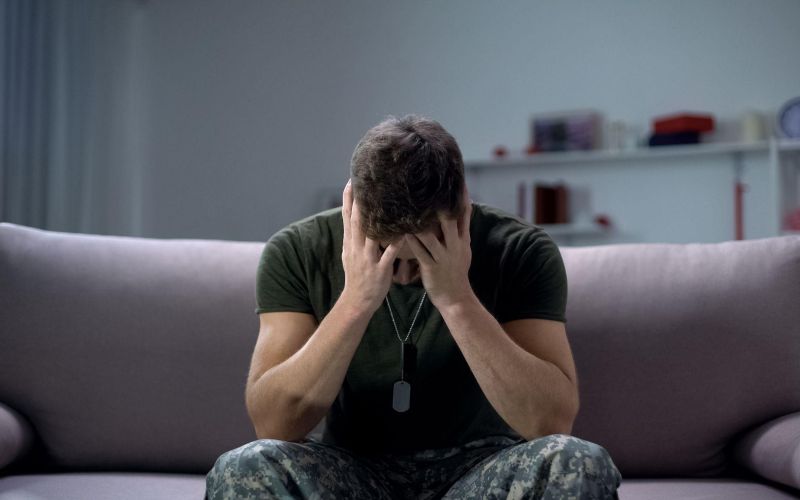 Man in military uniform sitting on sofa, hands covering face, representing stress or anxiety in a domestic setting.