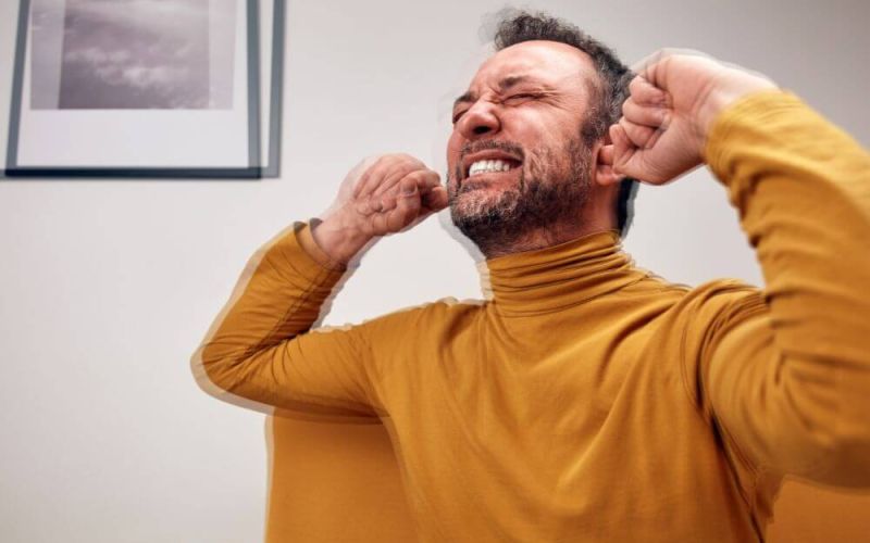 A man in a yellow turtleneck sits on a couch, grimacing and covering his ears with his hands, clearly distressed. A black-and-white photo hangs on the wall behind him.