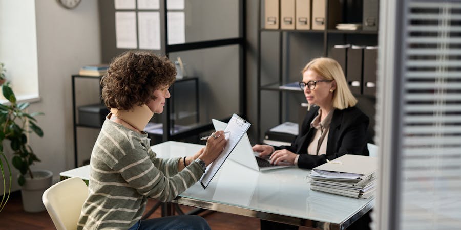 A person with curly hair sits at a glass table, writing notes with a pen, while another person in professional attire types on a laptop across from them in an office setting.