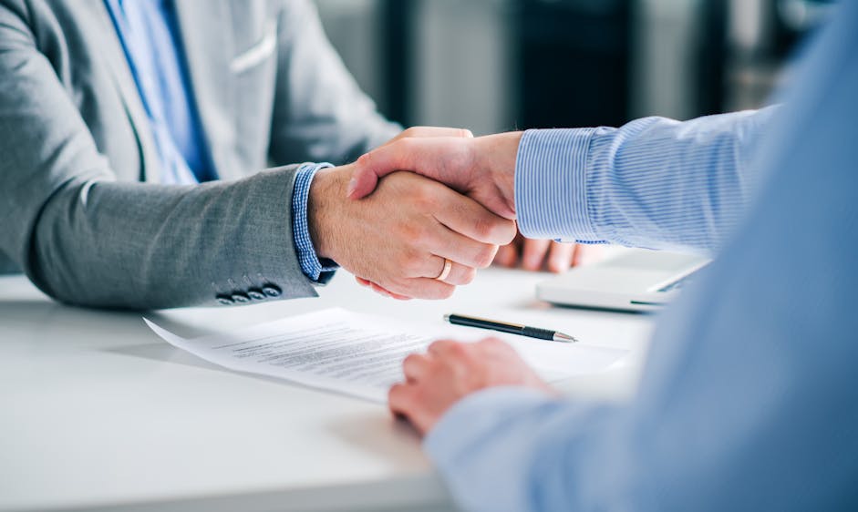 Two individuals are shaking hands over a desk, signifying agreement or greeting, with documents and a pen lying between them, likely in a professional setting.