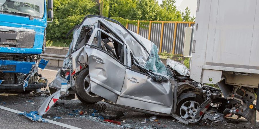 A catastrophic truck accident in Newport News, Virginia, showing a heavily damaged silver car crushed between a blue truck and a white truck. The car's structure is severely compromised, and debris is scattered across the road. Trees and a fence are visible in the background