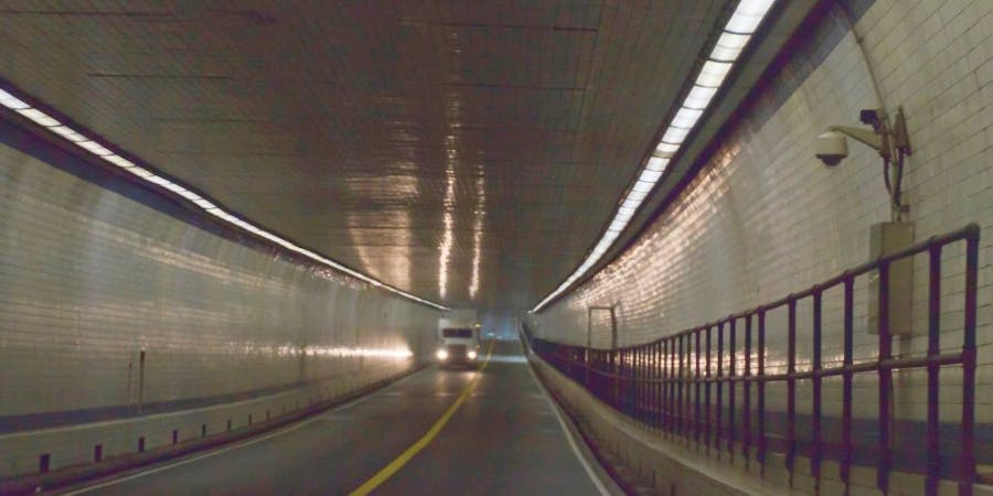 Interior view of the Hampton Roads Bridge-Tunnel showing a well-lit passage with reflective tiles on the walls. A truck is approaching from the opposite direction, driving through the tunnel, which has a road with clear lane markings and a metal railing along the side.