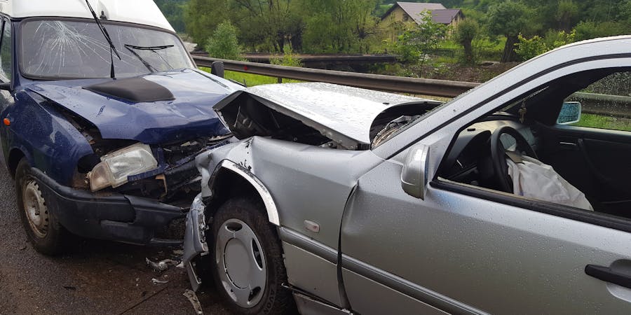 A close-up image of a head-on collision between a dark blue van and a silver sedan. The front ends of both vehicles are severely damaged, with the van's windshield shattered and the sedan's hood crumpled. The scene is set on a wet road with greenery and a house in the background, indicating a rural area. The airbags in the sedan are deployed, and debris is scattered around the crash site.
