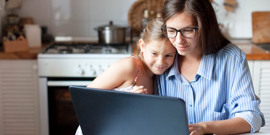 A woman and a young girl, embracing, are looking at a laptop in a home kitchen setting.