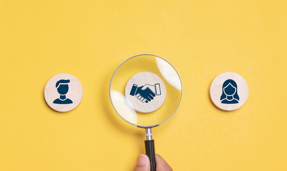 A magnifying glass is held over a handshake icon between two gender silhouettes on yellow background, suggesting scrutiny or focus on relationships or agreements.