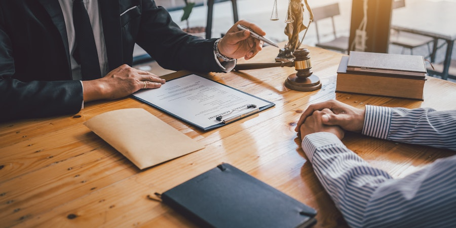 Two individuals in a meeting, one gesturing with hands over a legal document, in an office with law books and a justice scale. The context implies a legal consultation.