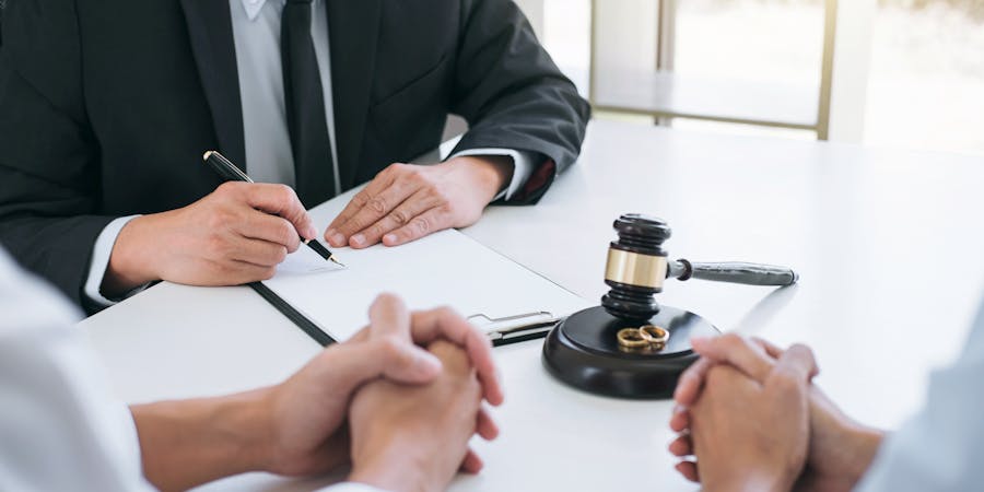 A person in a suit signs documents; another observes, near a gavel and rings on a table, suggesting a legal or matrimonial context.