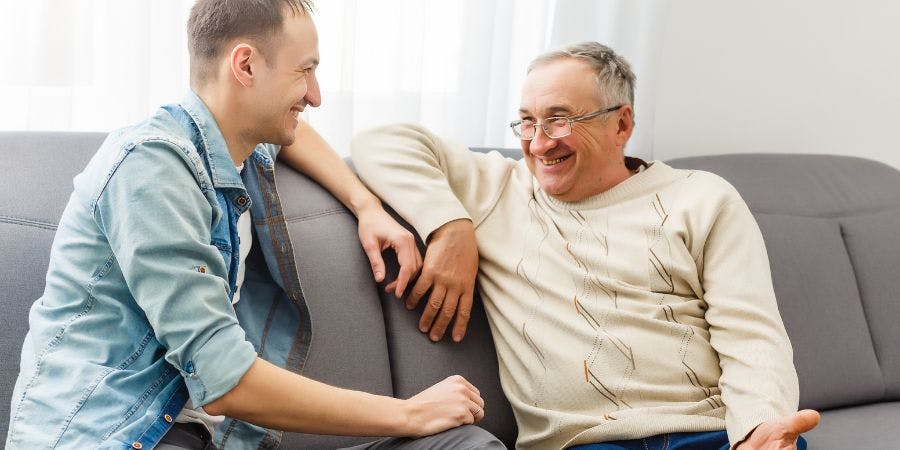 Two men are smiling and sitting on a couch, appearing to enjoy a pleasant conversation in a bright, indoor setting.
