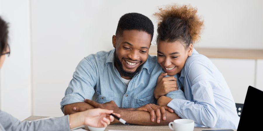 Two smiling people huddle, observing something with interest, likely on a table or desk, in a clean indoor setting with natural light.