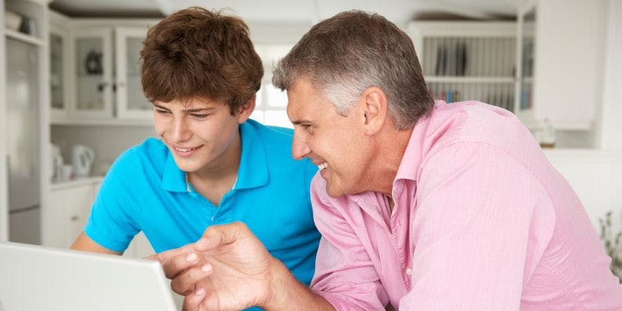 Two people, likely family, are smiling and looking at a laptop screen together in a brightly lit, modern kitchen.
