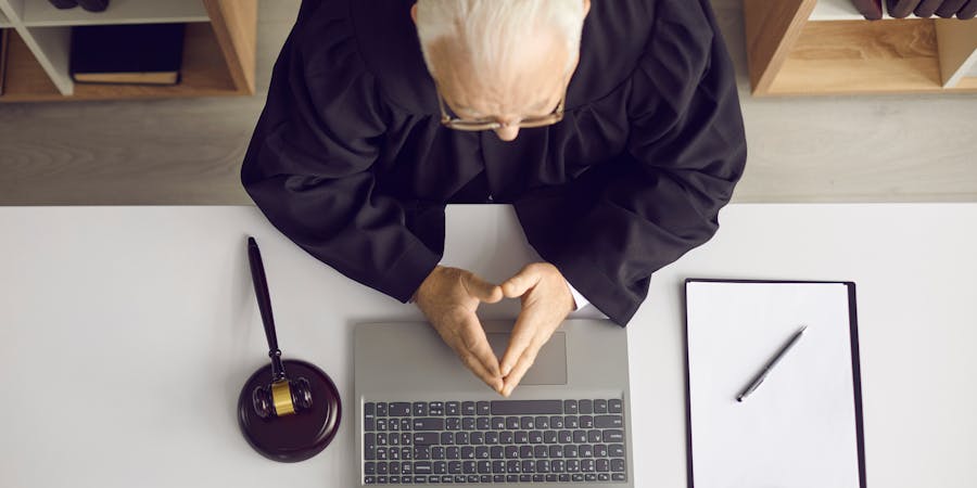 A judge in a black robe is working on a laptop at a white desk, beside a gavel and a notepad with a pen, signifying a legal context.