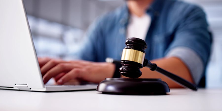 A gavel rests on a desk alongside a laptop. A person is typing on the laptop, suggesting engagement in legal work or research in a professional setting.