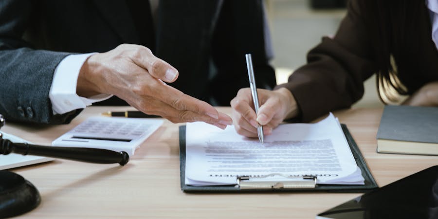 A hand gestures towards a document while another hand writes on it with a pen. The setting appears to be an office or meeting room, with a gavel and calculator nearby.