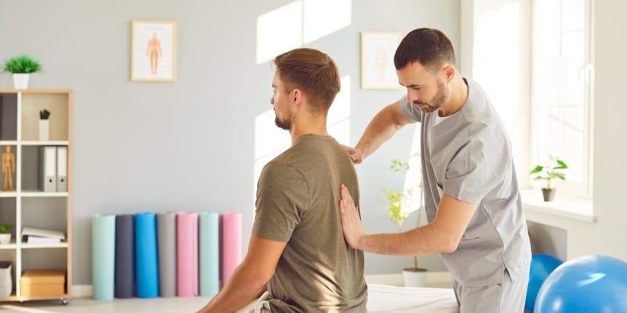 A male therapist in scrubs is assisting a seated male patient by applying pressure to his back. The therapy room features plants, exercise mats in soft colors, and large windows.