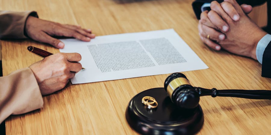 A hand holds a pen, signing a document on a wooden table. A gavel and two wedding rings sit nearby, suggesting a legal or marital context. Another set of hands rests on the table.