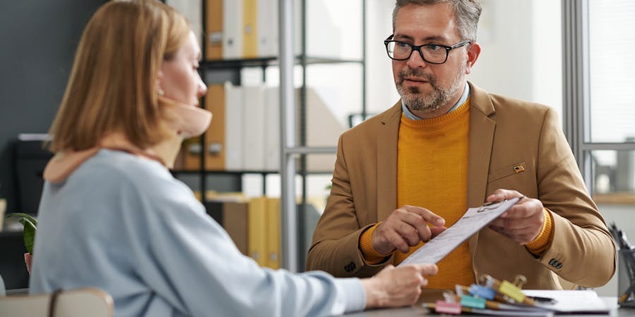 A man in a brown blazer and orange sweater hands a document to a woman in a light blue sweater during a discussion at a modern office table filled with colorful stationery.