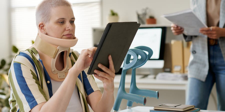A woman wearing a neck brace sits at a desk, using a tablet. Another person stands nearby holding papers in an office setting with visible furniture and a computer in the background.